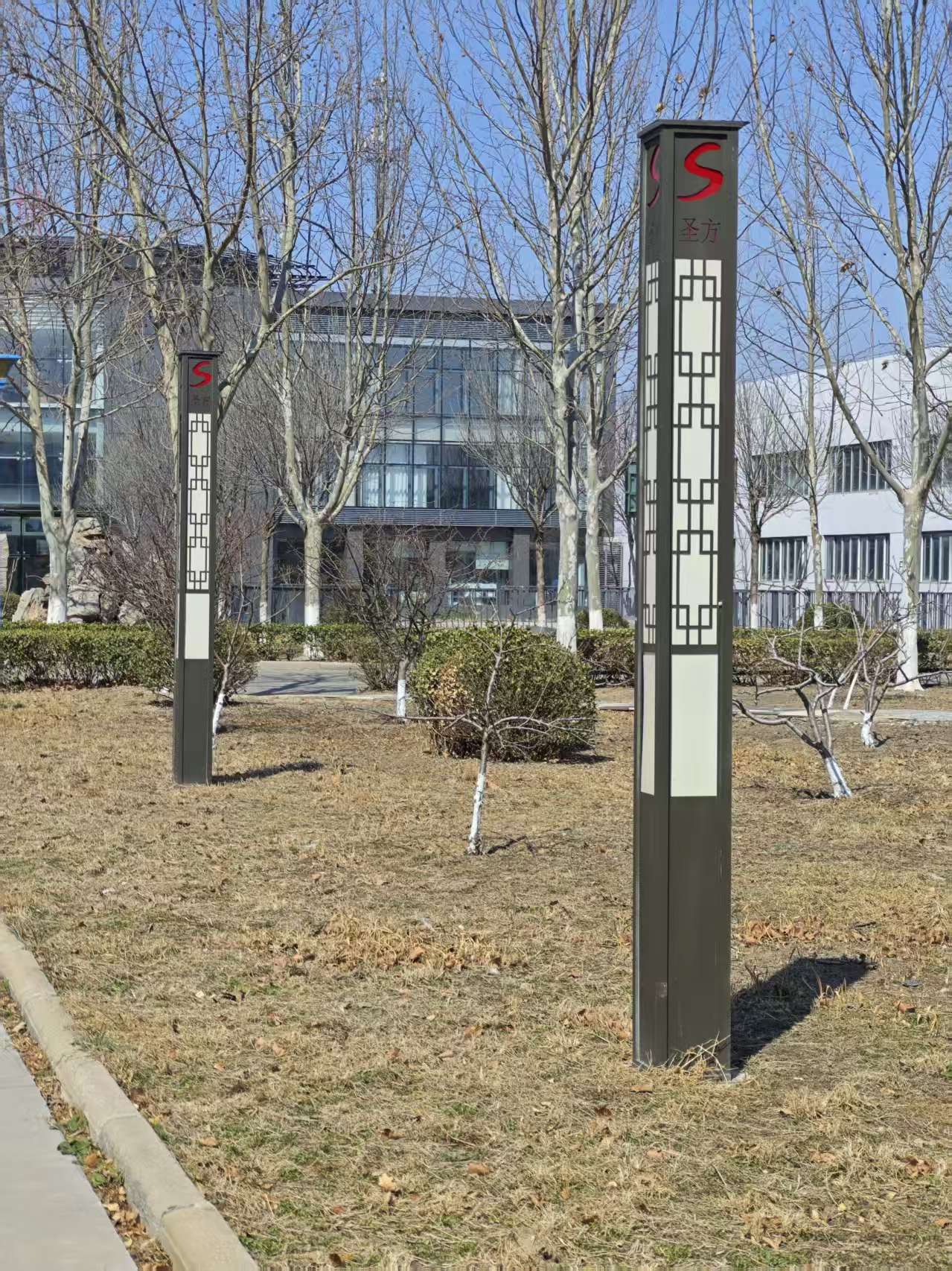 这是一组新中式景观灯柱，深灰色立柱搭配中式回纹镂空格栅，顶部点缀红色 “S” 标识与 “圣方” 字样，简约大气，既保留了传统窗棂的东方韵味，又兼具现代户外照明的实用功能，与园区的建筑、植被环境自然融合。