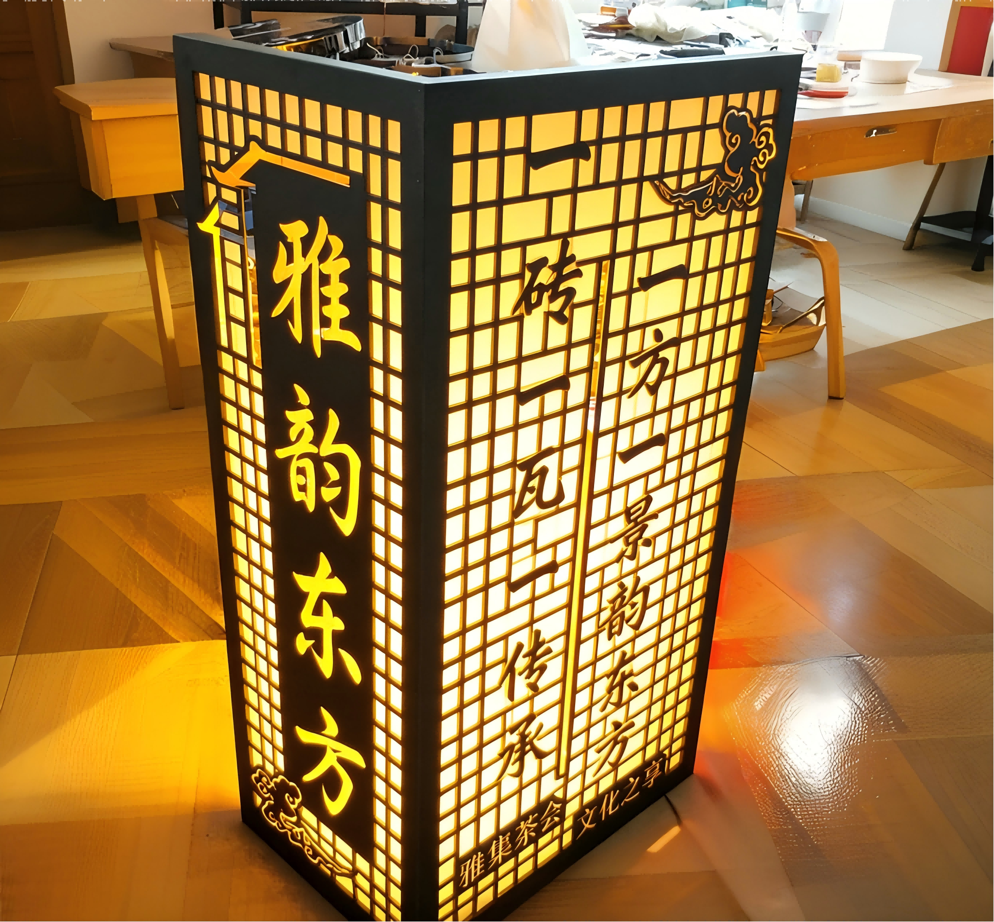 这是一盏新中式风格的立式镂空发光灯箱，整体呈方正立柱造型，以沉稳的黑色金属或木质框架勾勒出精致的格栅纹理，暖黄色灯光从镂空处透出，营造出温润雅致的氛围感。